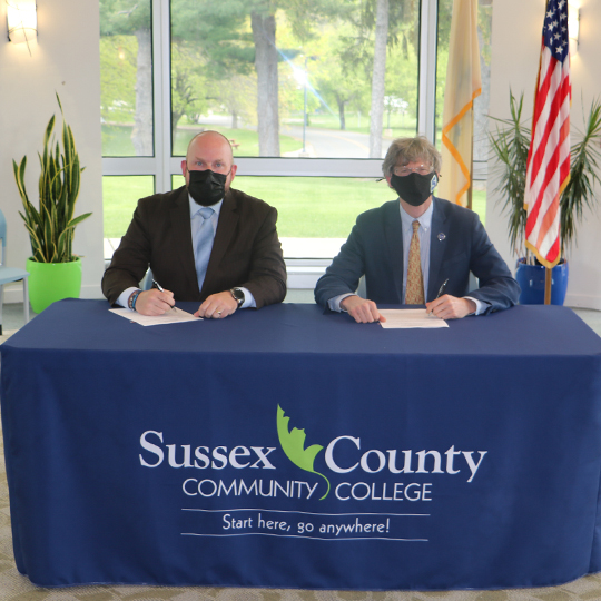 2 men in suits sit at a table with a blue tablecloth and college logo signing an agreement