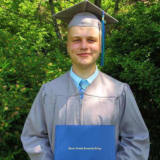 Young, white male standing outside with his gray graduation cap and gown holding his diploma.