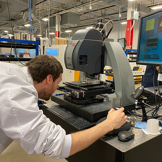 A male student in a lab coat works on machinery in the Optics technology program.