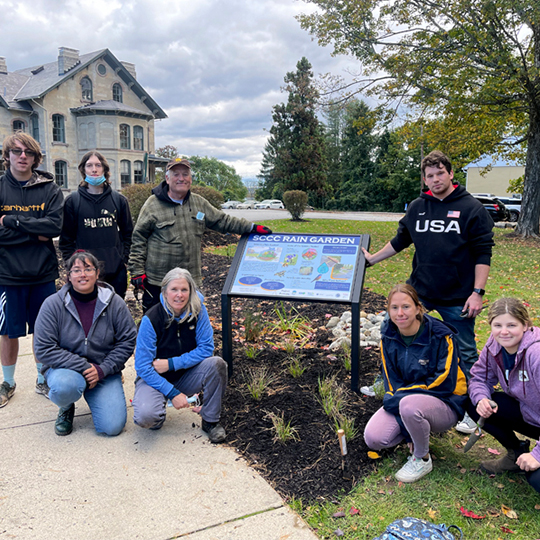 Students pose outside near a sign and the plantings from the rain garden.
