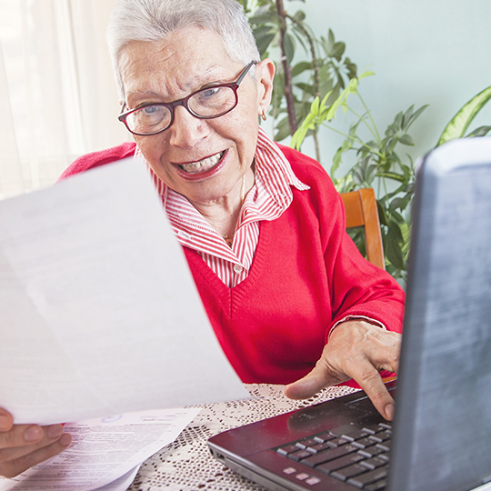 Image of an elderly woman wearing a red shirt on a laptop holding a paper in her hand.