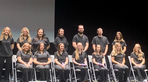 14  nursing students stand and are seating for a photo on stage.