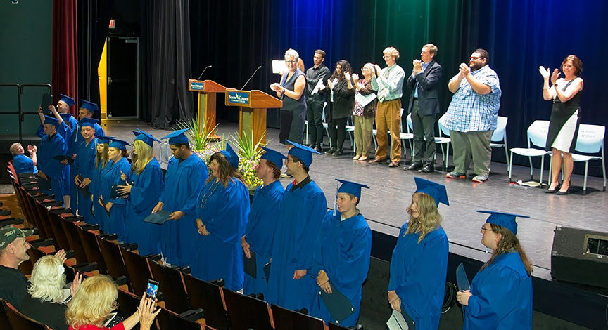 Graduates in blue cap and gown face the crowd for accolades.